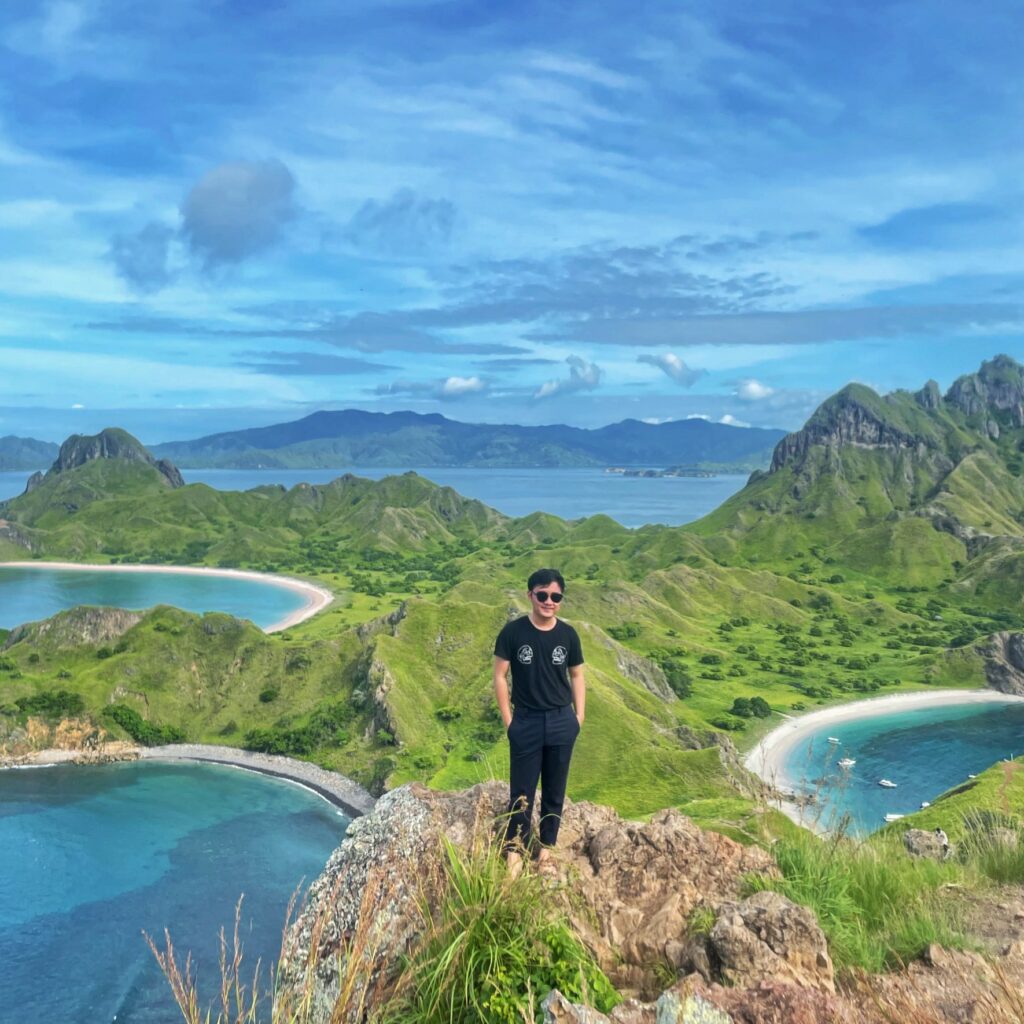 The picture shows Kevin visiting Labuan Bajo. He is standing on a cliff and behind him there are mountains and water.