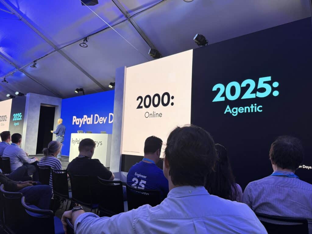 An audience view of a PayPal Dev Days presentation. The stage shows large display panels with text comparing eras: '2000: Online' and '2025: Agentic'. A presenter in a blue shirt stands at a podium labeled 'PayPal Dev Days'. The audience consists of seated attendees wearing casual business attire, with one visible person wearing a blue shirt with 'Engineer 25' on the back. The venue has overhead lighting and appears to be in a large tent or temporary structure with a white ceiling.