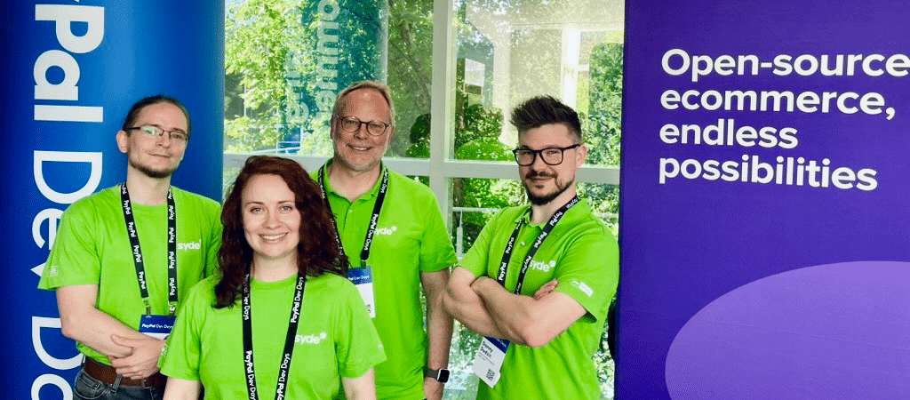 A group of four people wearing bright green t-shirts and lanyards standing at what appears to be a conference or event booth. They are positioned between two promotional banners - a blue one reading 'PayPal Dev Days' on the left and a purple one for WooCommerce on the right that says 'Open-source ecommerce, endless possibilities.' The group members are wearing matching conference badges and standing in front of large windows with greenery visible outside.