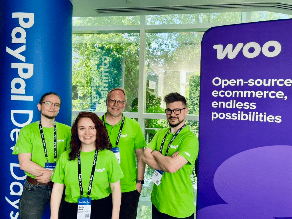 A group of four people wearing bright green t-shirts and lanyards standing at what appears to be a conference or event booth. They are positioned between two promotional banners - a blue one reading 'PayPal Dev Days' on the left and a purple one for WooCommerce on the right that says 'Open-source ecommerce, endless possibilities.' The group members are wearing matching conference badges and standing in front of large windows with greenery visible outside.