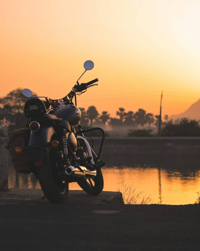 A matte black Royal Enfield Classic silhouetted against a warm golden sunset, parked beside a still lake with palm trees and mountains in the hazy background.