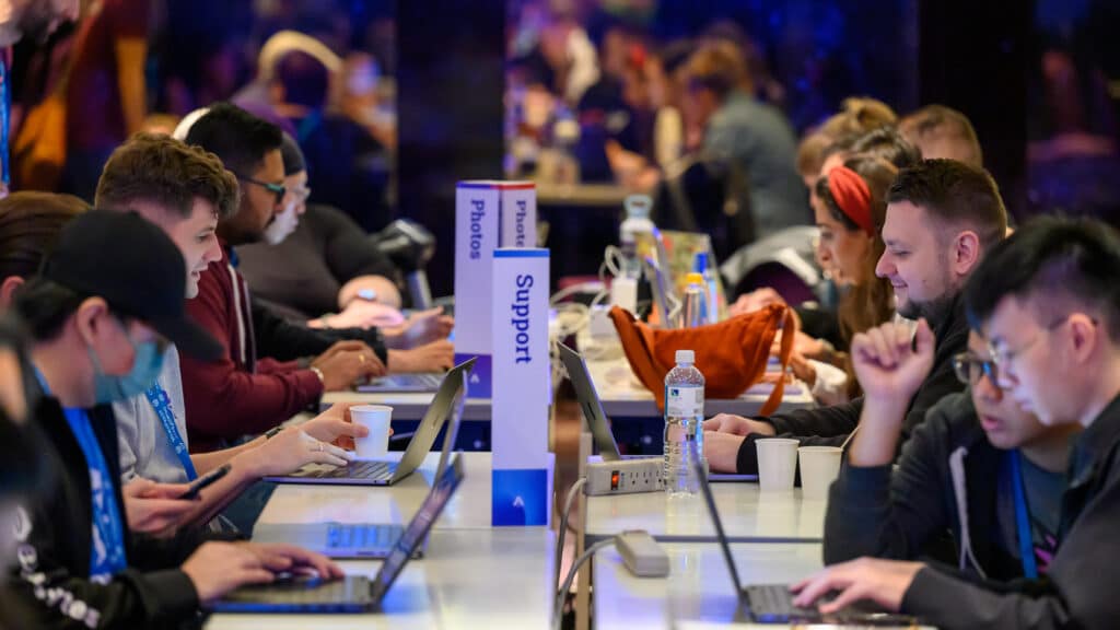 Attendees working on laptops at long tables during WordCamp Asia 2025 Contributor Day, with "Photos" and "Support" contribution table signs visible in the center, blue lanyards and a buzzing conference atmosphere in the background.
