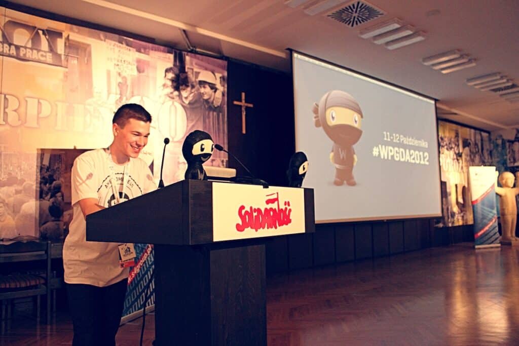 A younger Danny at a Solidarność-branded podium at WordCamp Gdańsk 2012, with WordPress ninja plush toys on the lectern and the #WPGDA2012 slide projected on screen behind.